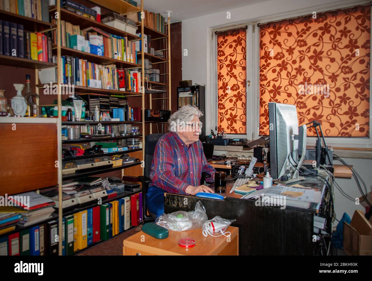 Elderly white haired man working at home on the desktop computer in a mess. Bookshelf with many books and antiviral masks ready Stock Photo