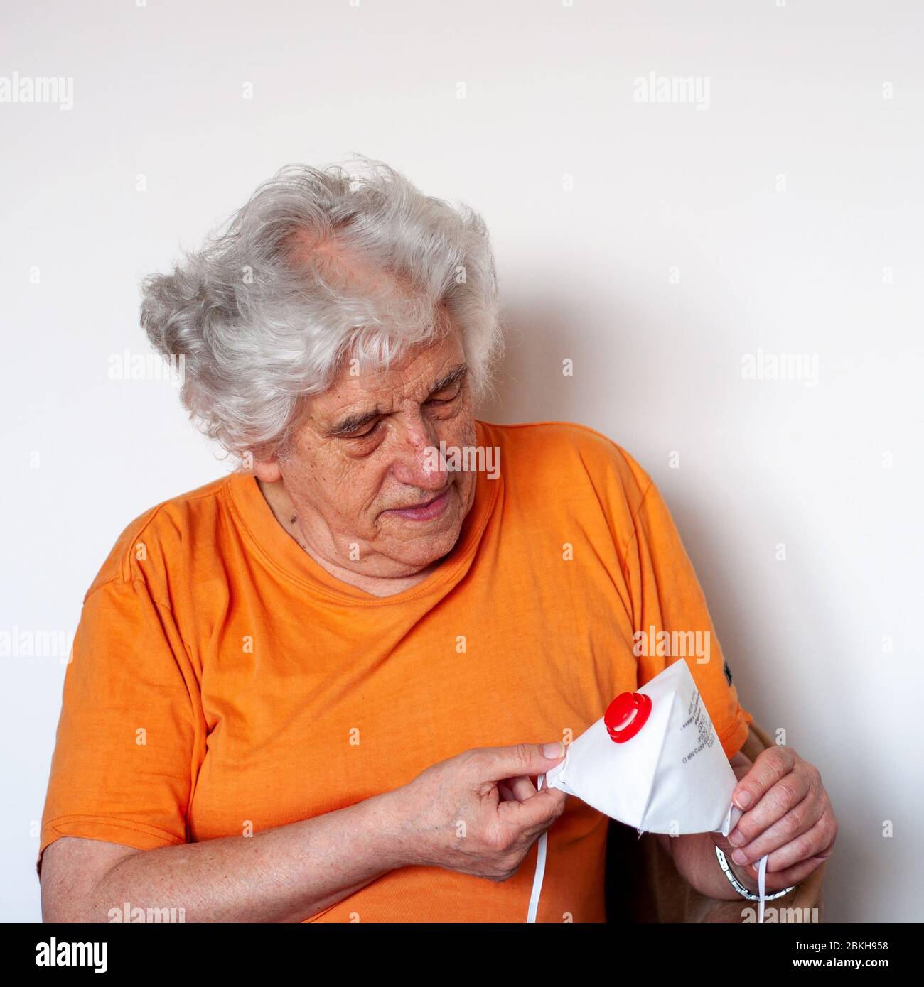 A senior studying  an antiviral protective FFP3 N99 mask, protecting against coronavirus. White haired Caucasian man in orange T-shirt on white wall Stock Photo