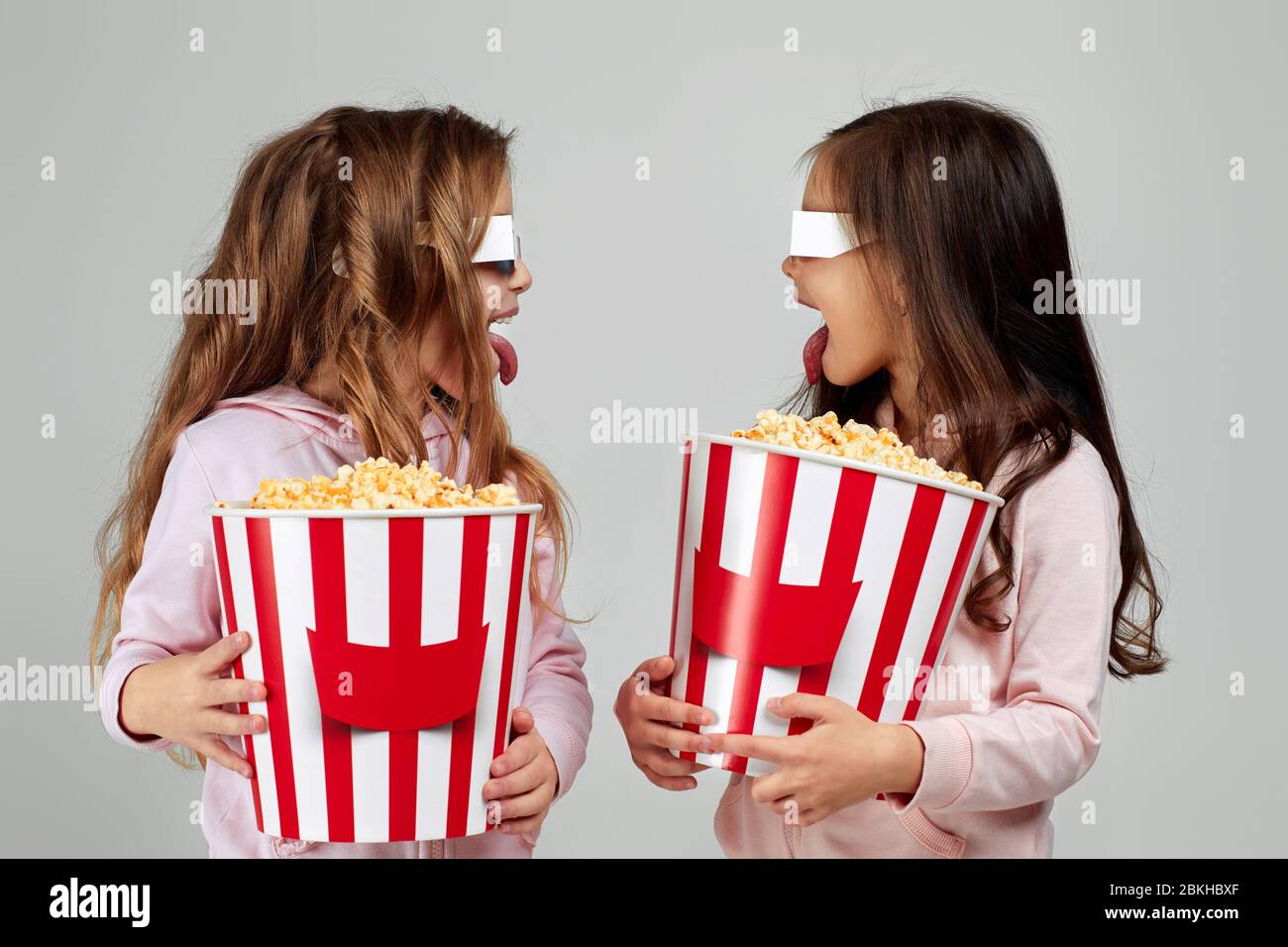 two funny caucasian smiling little girls in red-blue 3d glasses holding popcorn buckets and and looking at each other, close-up. kids showing tongue Stock Photo