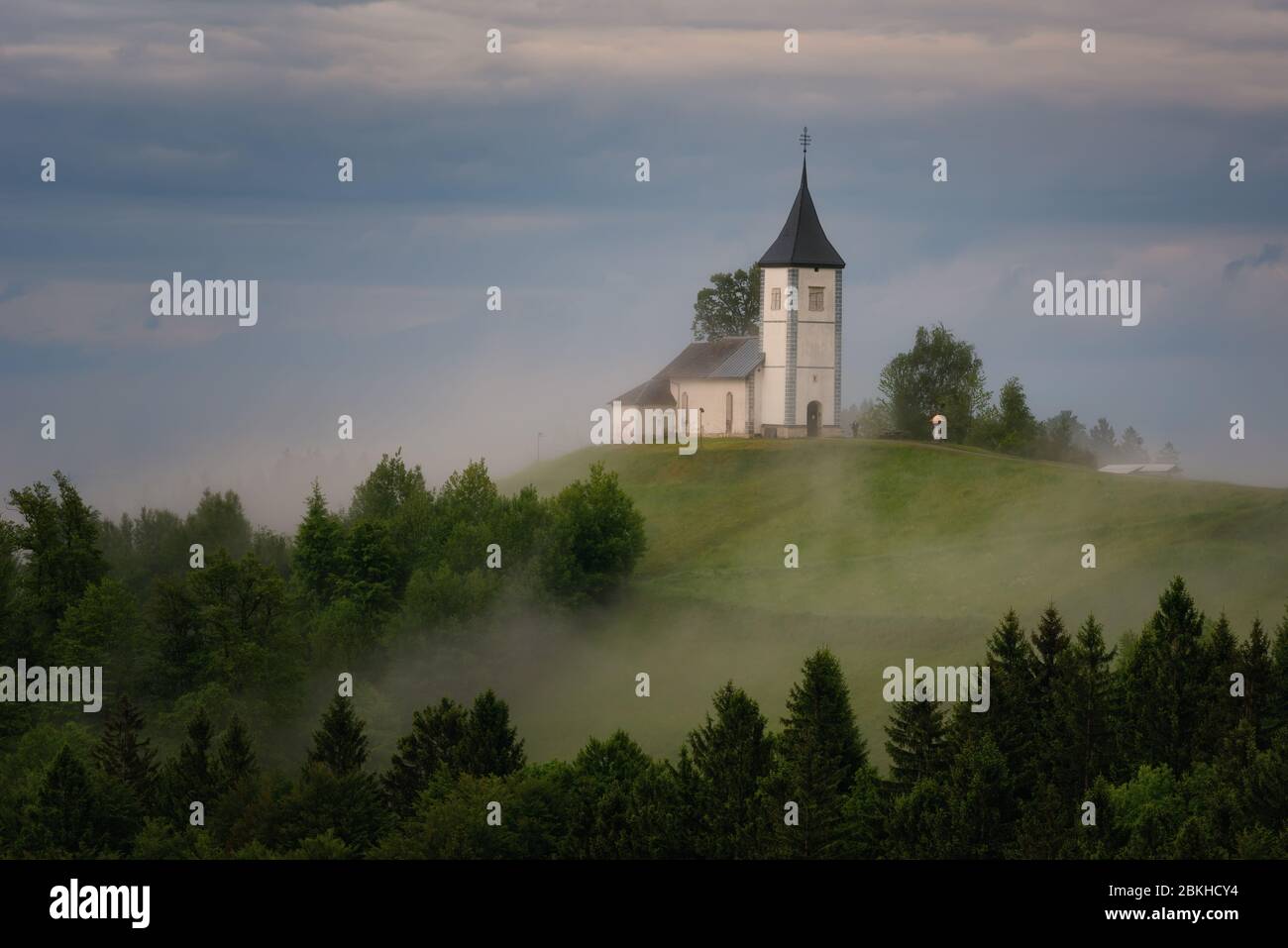 Jamnik church on a hillside in the spring, foggy weather at sunset in Slovenia, Europe. Mountain landscape shortly after spring rain. Slovenian Alps. Stock Photo