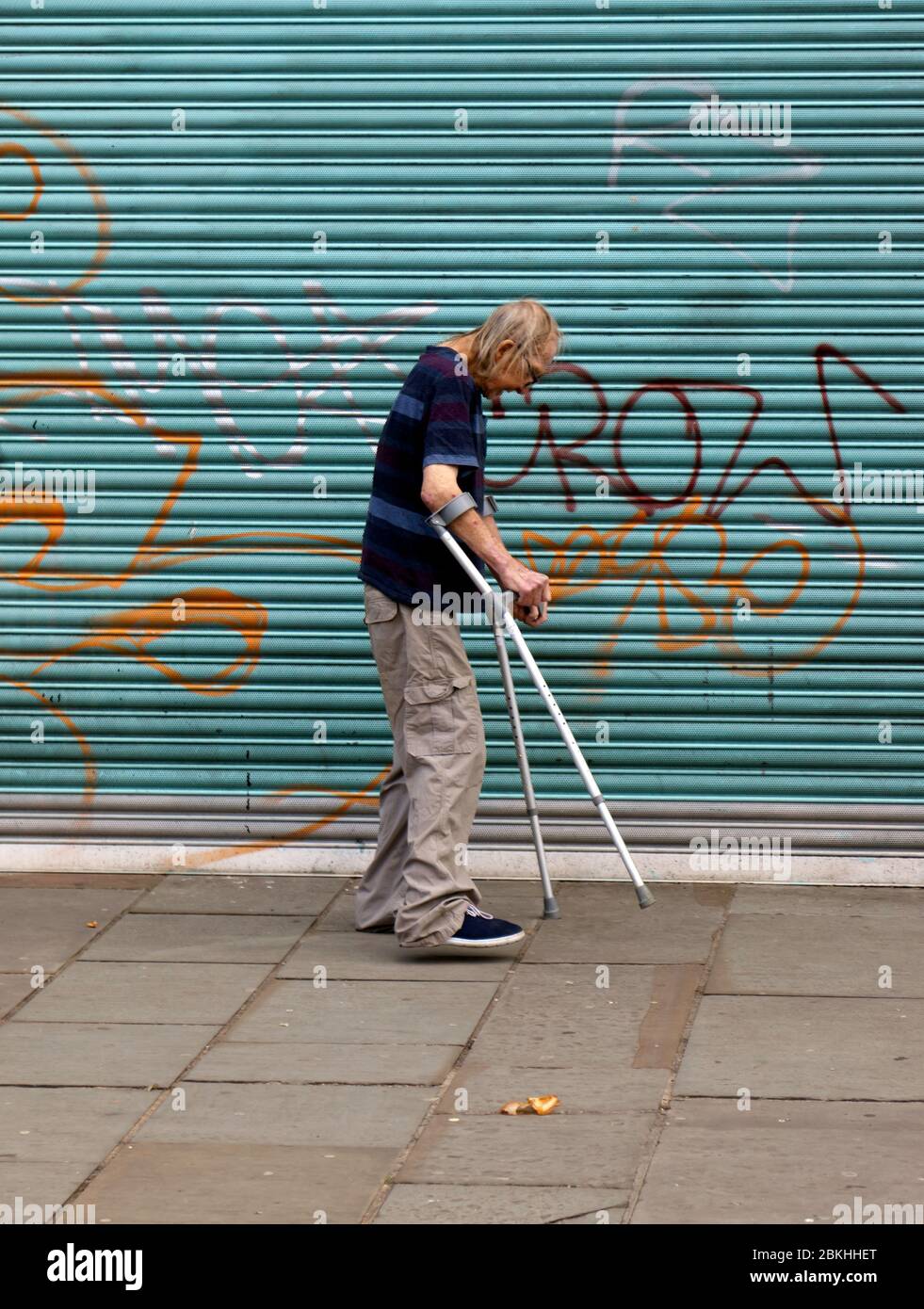 A vulnerable, senior Man, using crutches, makes his way slowly down Lewisham High Street past shops which are currently forced to closed due to  the 2019-2020 COVID-19 Pandemic Stock Photo