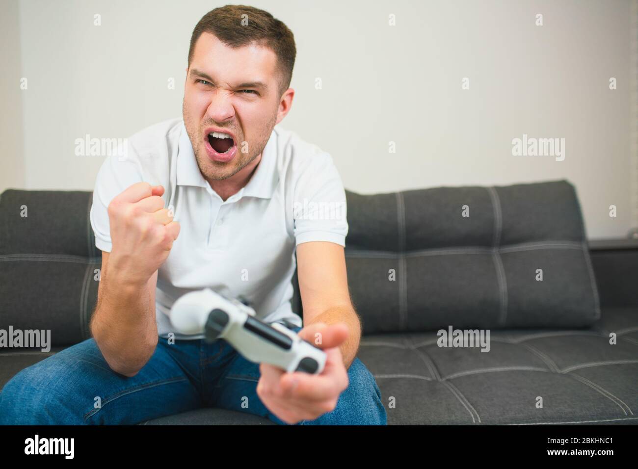 Young man sit in room during quarantine. Emotional guy playing computer games. Male person hold joystick in hand and cheer with pleasure. Screaming Stock Photo
