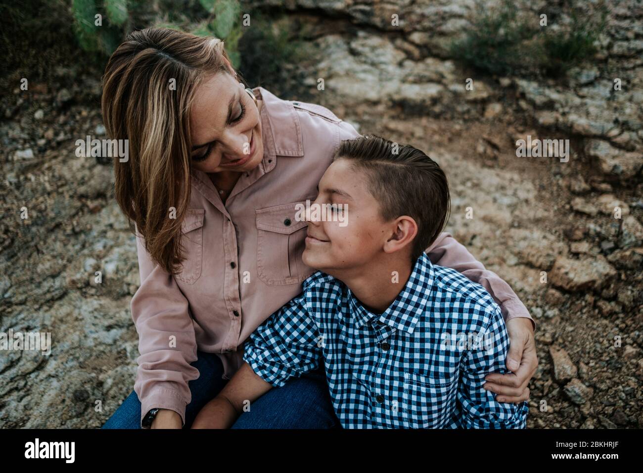 Mother hugging non binary child while sitting on rocks Stock Photo