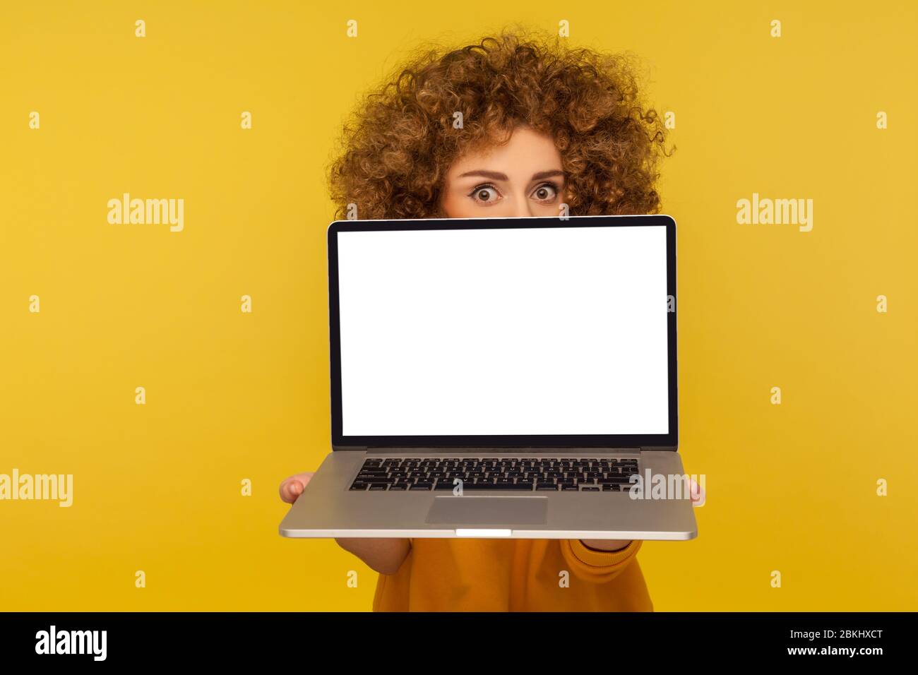 Scared shocked woman with curly hair peeking from behind laptop blank screen with frightened worry eyes, mock up display for cybersecurity advertiseme Stock Photo