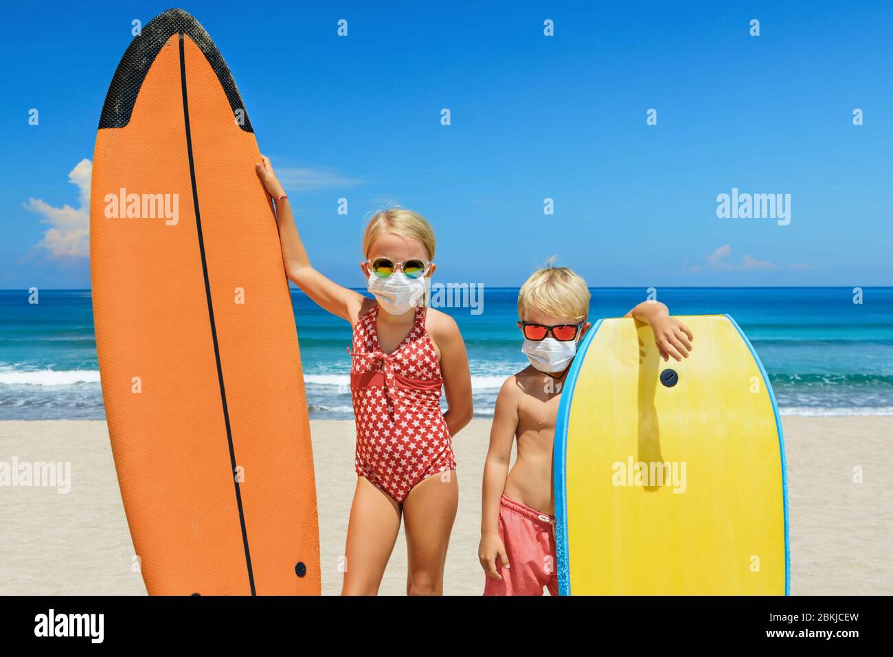 Funny kids with surf boards wear protective mask on sea beach. Cancelled cruises, tours due coronavirus COVID 19 world epidemic. Stock Photo