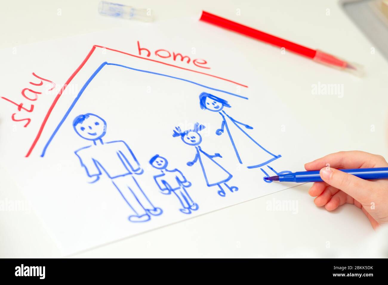 Child is drawing happy family with words Stay Home on white sheet of paper at home. Children creativity. Stock Photo