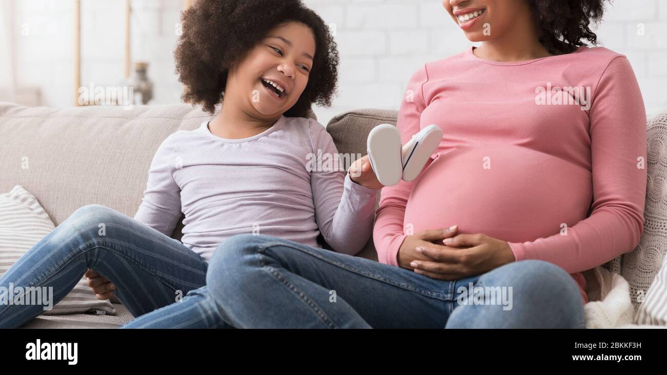 Joyful Little African Girl Playing With Baby Shoes Near Mom's Pregnant Belly Stock Photo