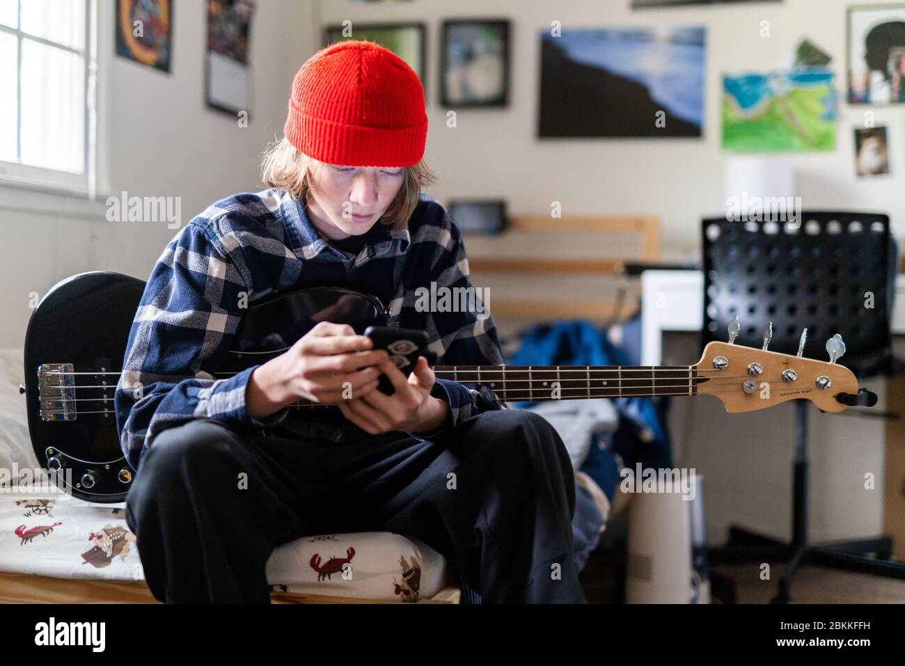 Teenager looking at smart phone while holding bass guitar in bedroom Stock Photo