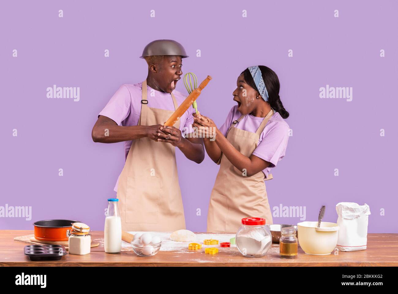 Emotional African American girl and guy fighting with kitchen utensils while cooking on color background Stock Photo