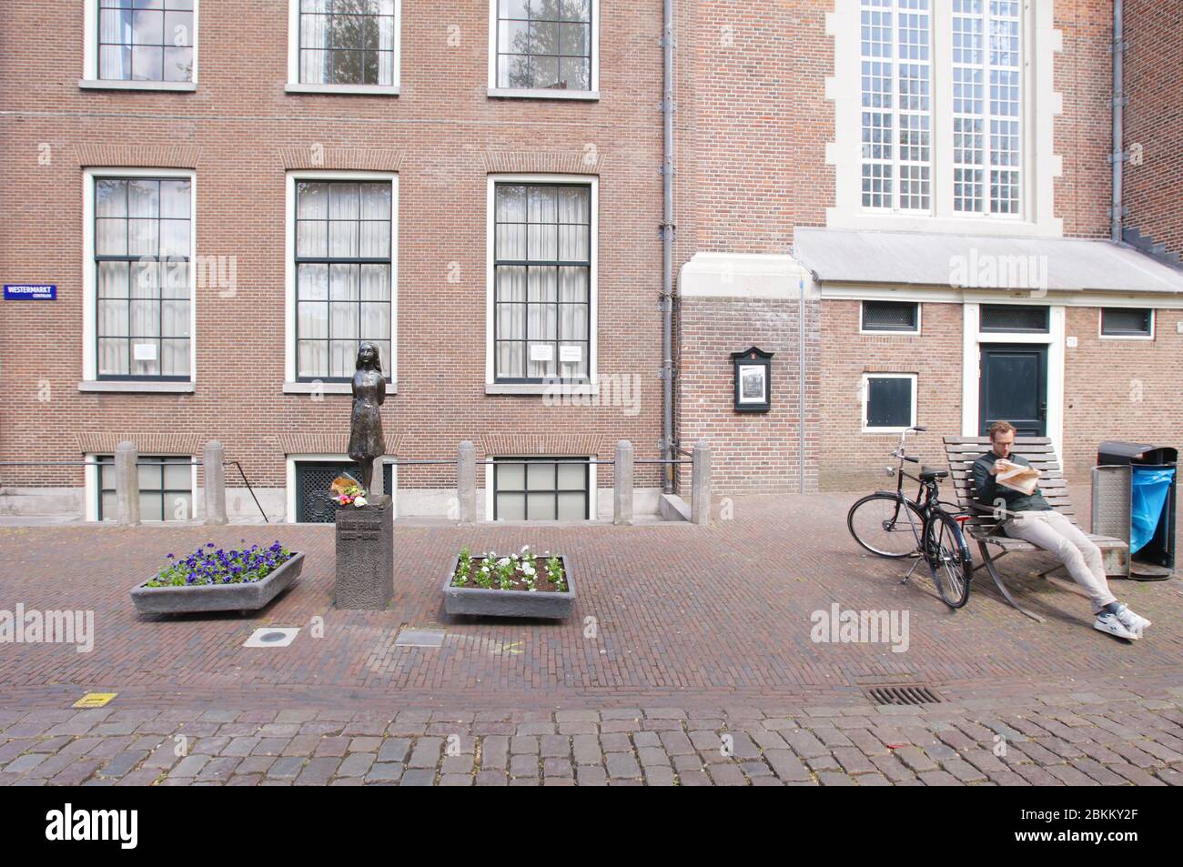 A local resident reading newspaper near flowers laid at the Anne Frank (1929-1945) statue near Anne Frank house on empty street during the Remembrance Stock Photo
