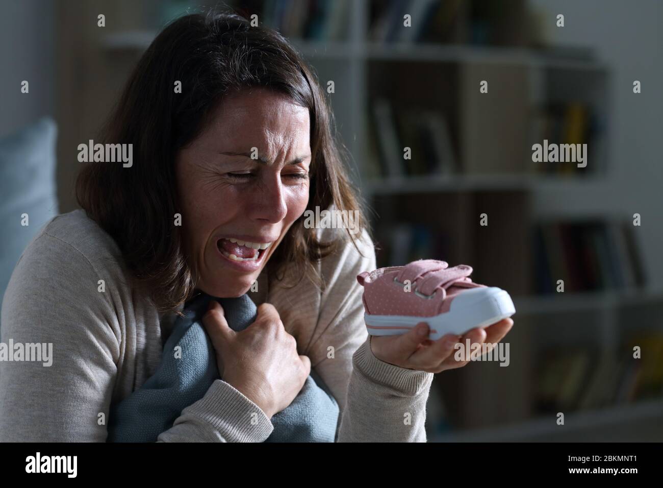 Sad mother crying missing her lost baby daughter holding shoe sitting at night at home Stock Photo