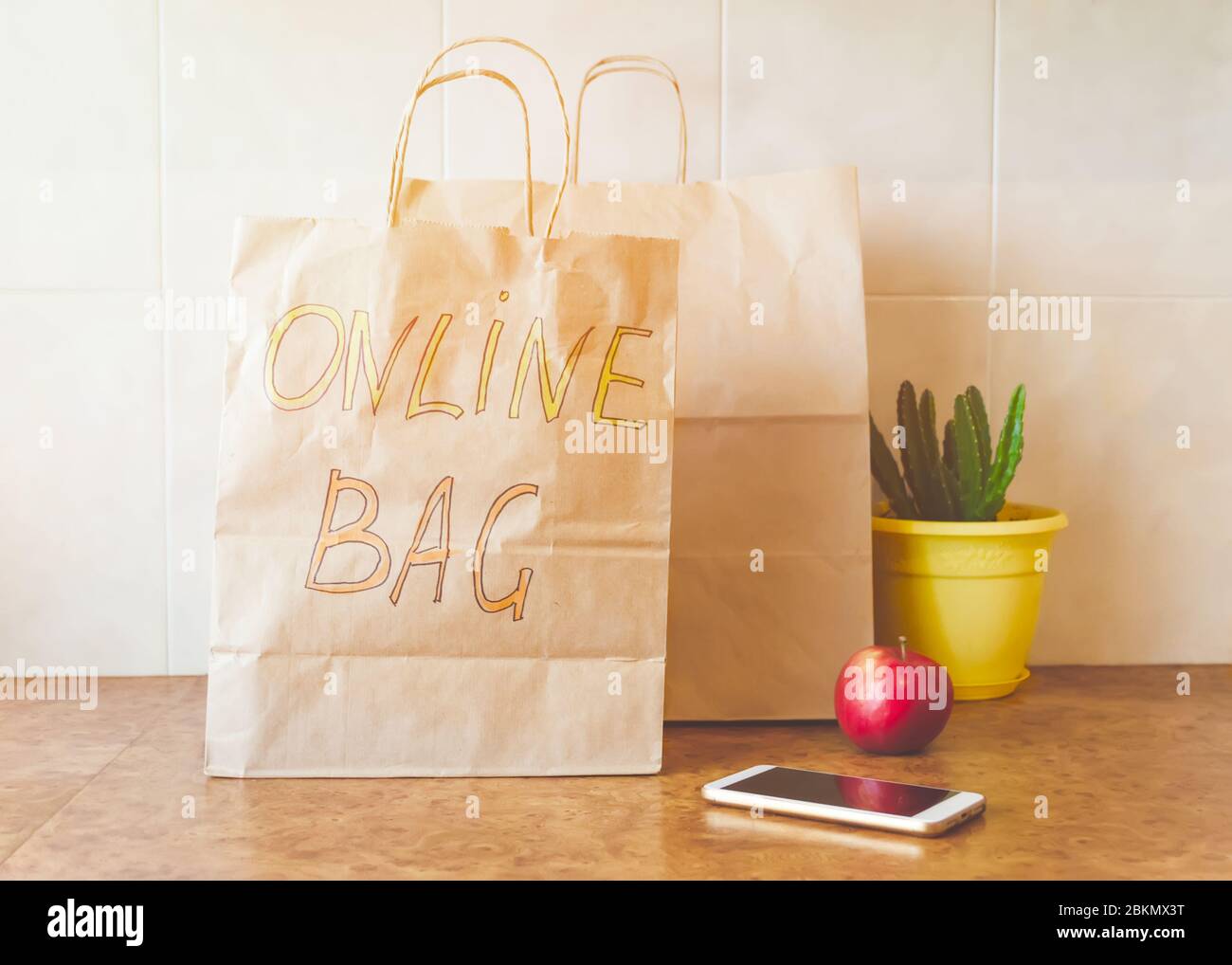 Paper bag with inscription - Online bag, fresh red apple, mobile phone and a houseplant in a yellow pot on a kitchen table. Online shopping and contac Stock Photo