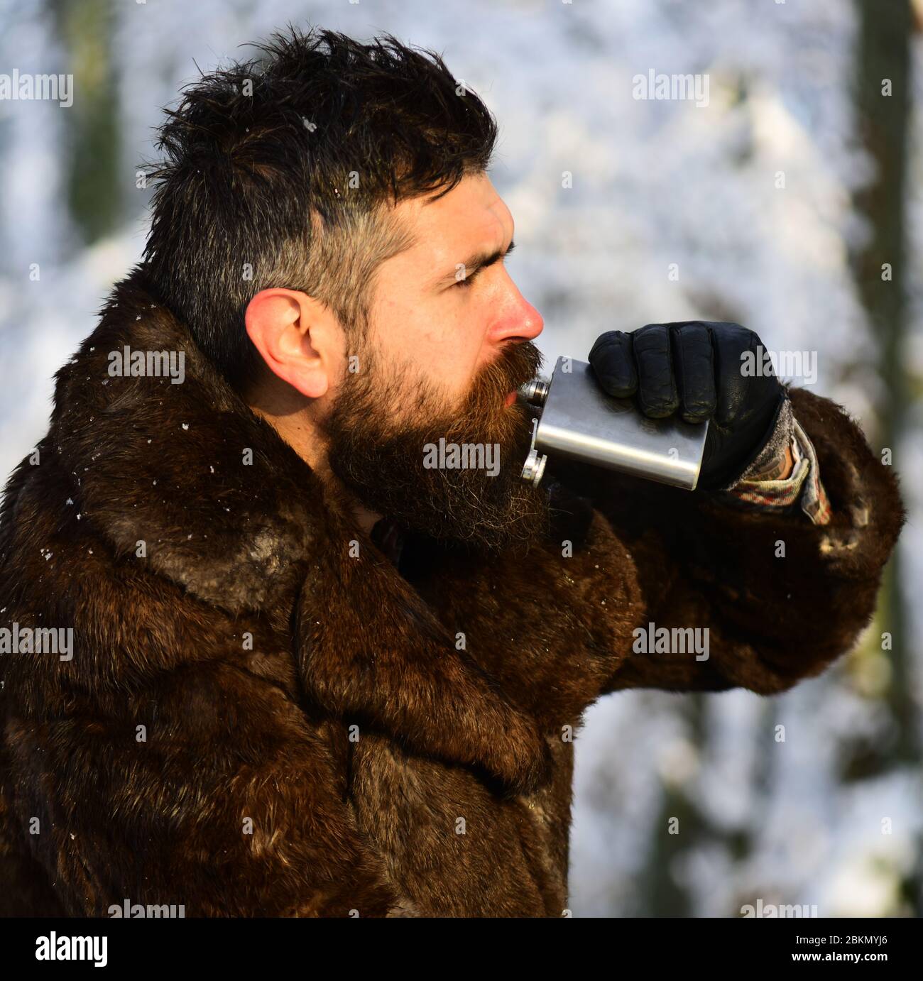 Guy on snowy nature park background, defocused. Warming drinks concept. Man in fur coat drinks from metal flask. Macho with beard and mustache in forest. Stock Photo