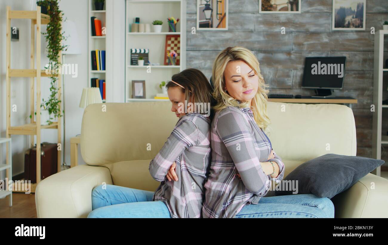 Little girl with bad behavior sitting back to back with her mother on the couch after a dispute. Stock Photo
