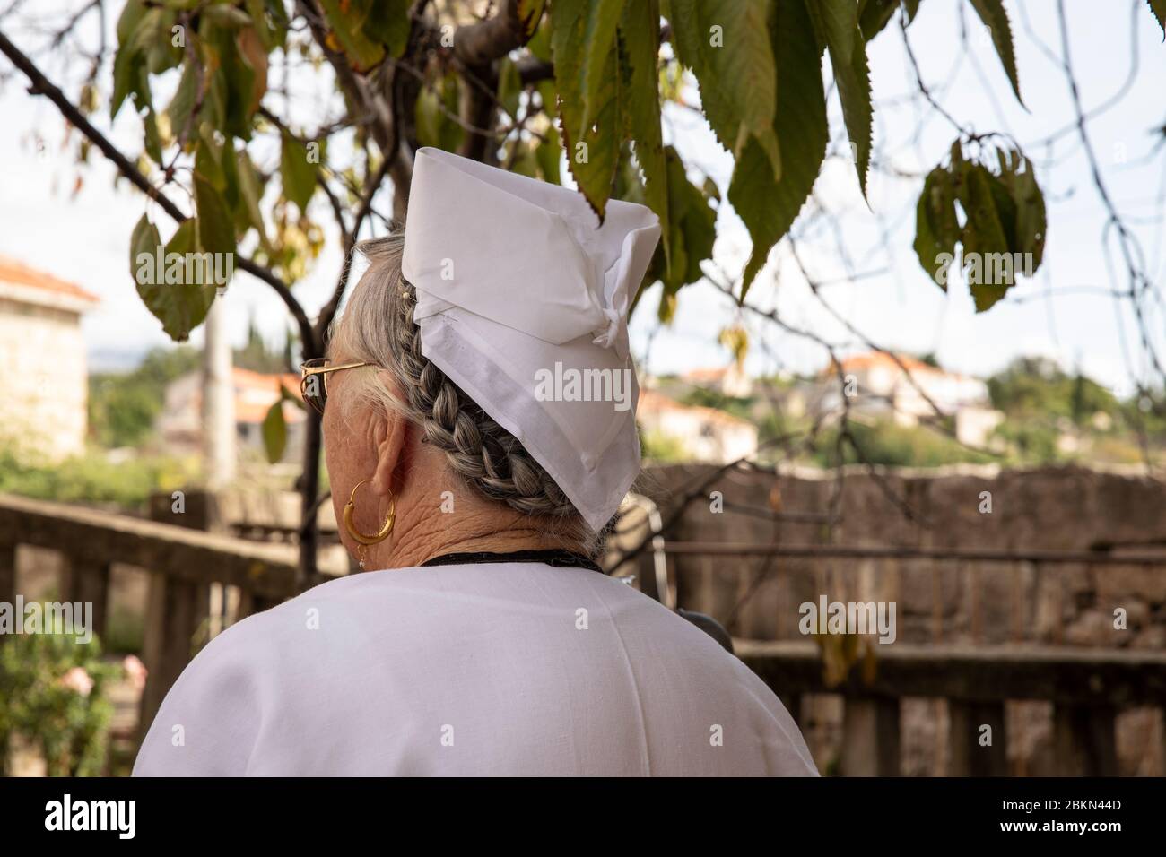 An elderly woman wearing typical Dalmatian golden earrings and a traditional folklore costume with a white kerhief hat and braided hair in Konavle, Da Stock Photo