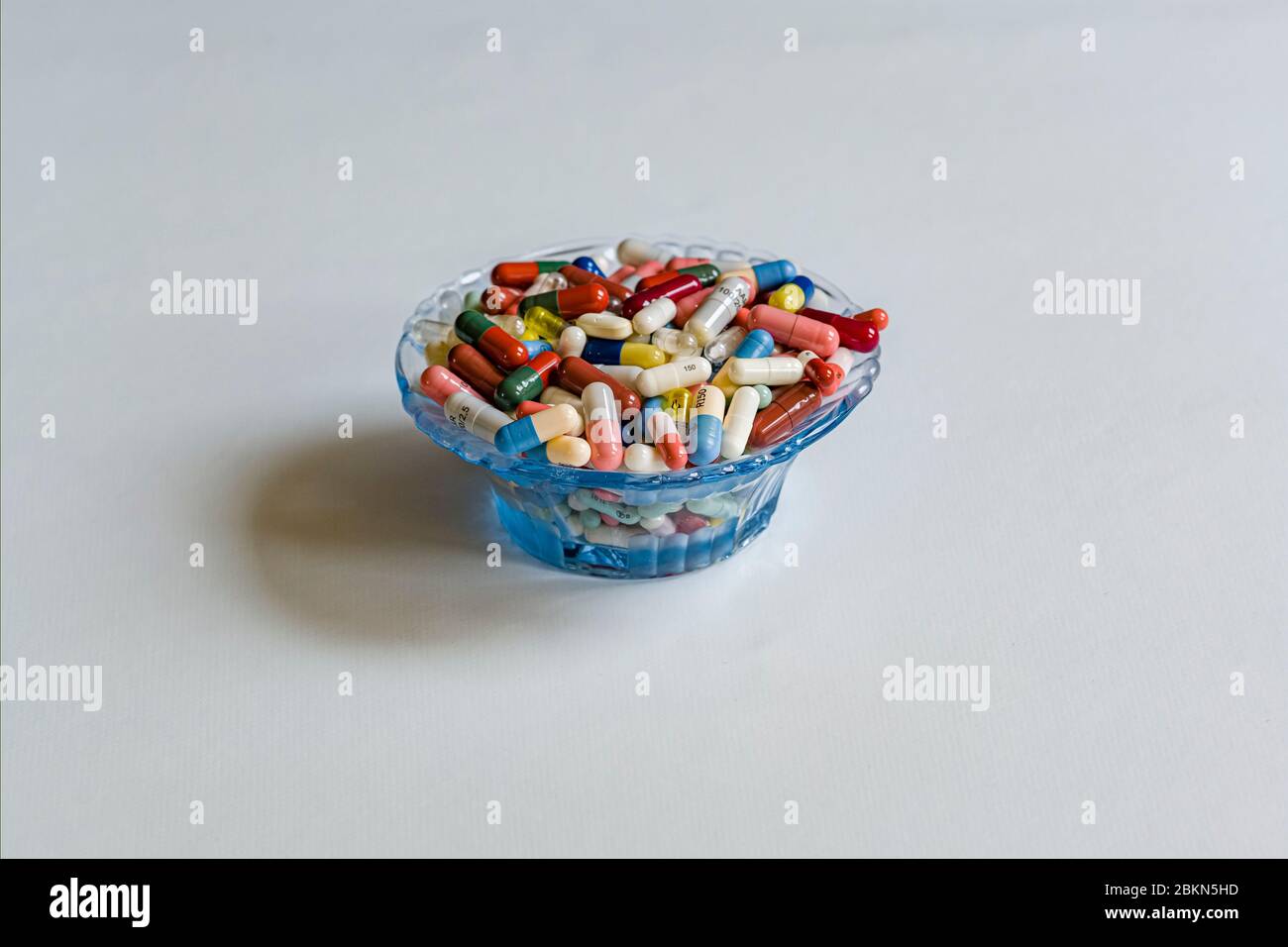 A small dish filled with differently colored medical capsules, displayed on a white table Stock Photo