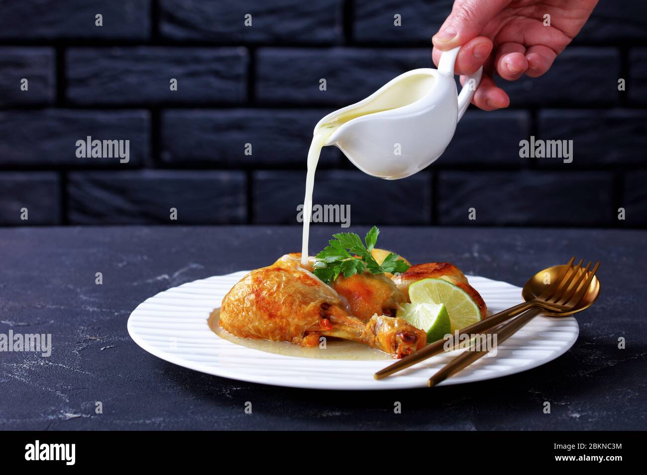 Woman hand is pouring dripping coconut sauce on top of roasted chicken leg served with baby potatoes on a white plate, horizontal view from above Stock Photo