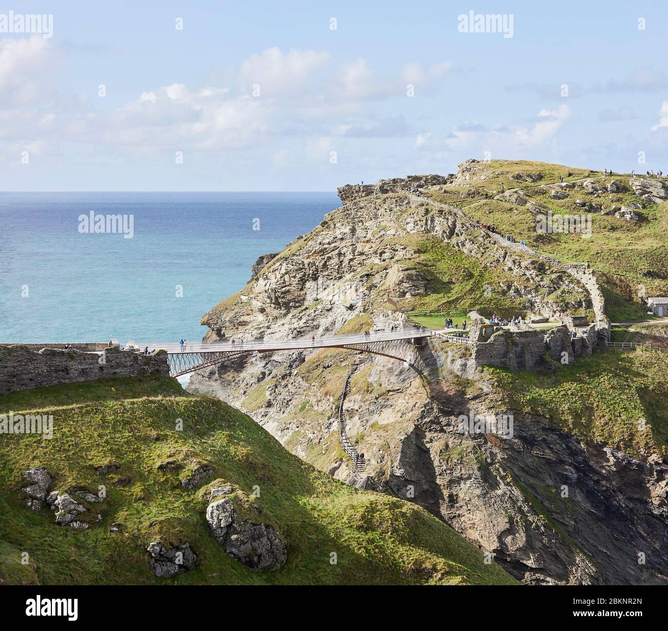 Distant view with visitors walking on bridge and medieval ruins of Tintagel Castle. Tintagel Bridge, Tintagel, United Kingdom. Architect: William Matt Stock Photo