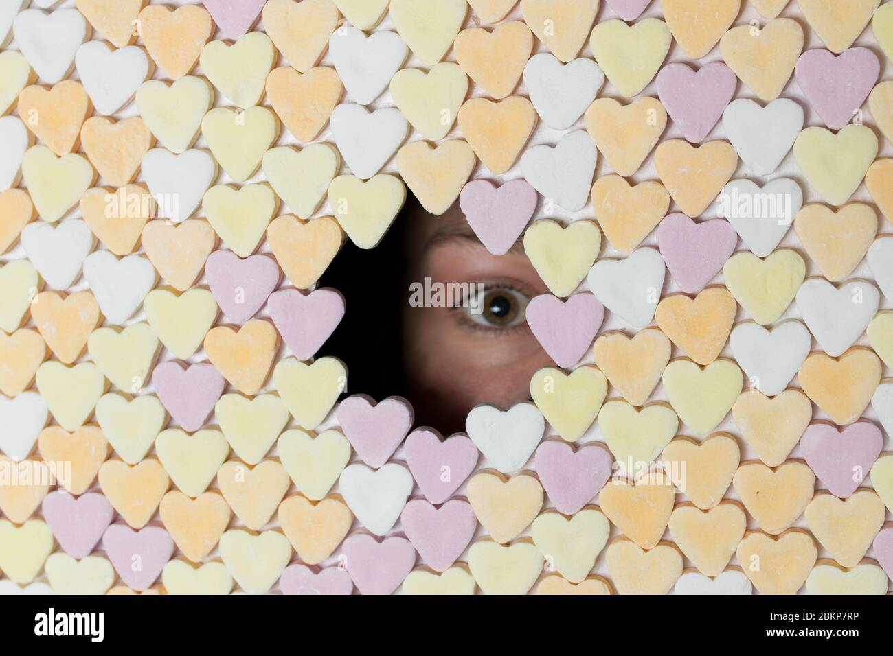 Girl peaking with one eye through hole in wall of heart-shaped sweets. Royalty free stock photo. Stock Photo