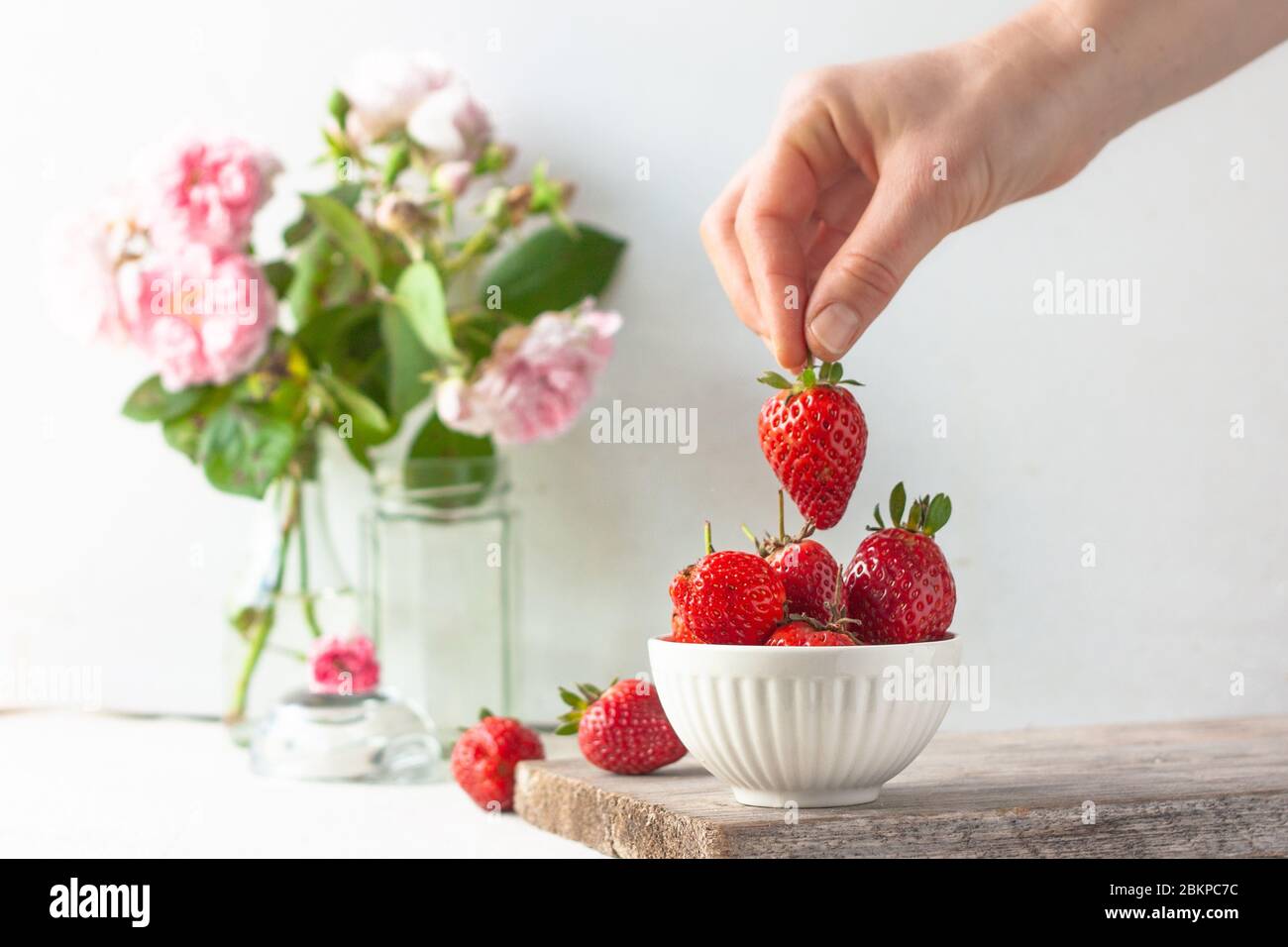 Woman takes or puts one strowberry in fruit bowl full of fresh, red, jucy strawberries. Pastel-colored decoration.  Stock Photo