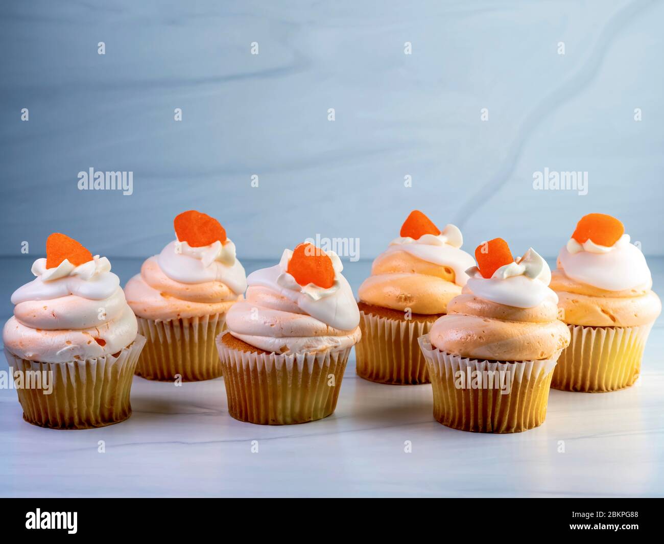 Six, 6, yellow cupcakes lined up with orange and white creamsicle frosting swirled up high and an orange gum drop candy on top.  Decadent delicious de Stock Photo
