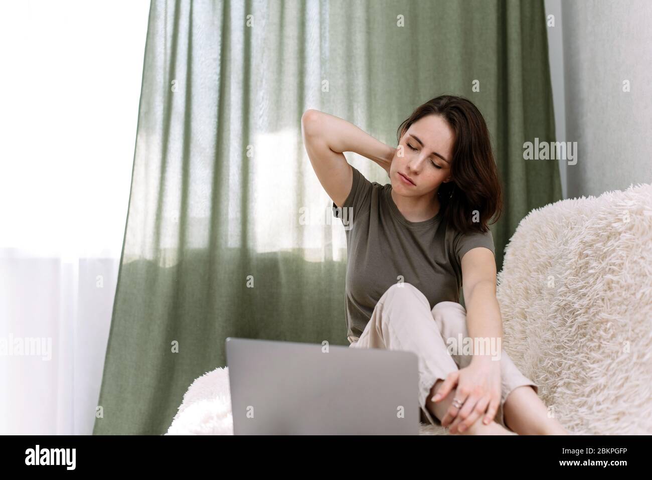 A woman works at home with a laptop and rubs her neck in pain. Pain in the neck from prolonged work at the computer. Stock Photo