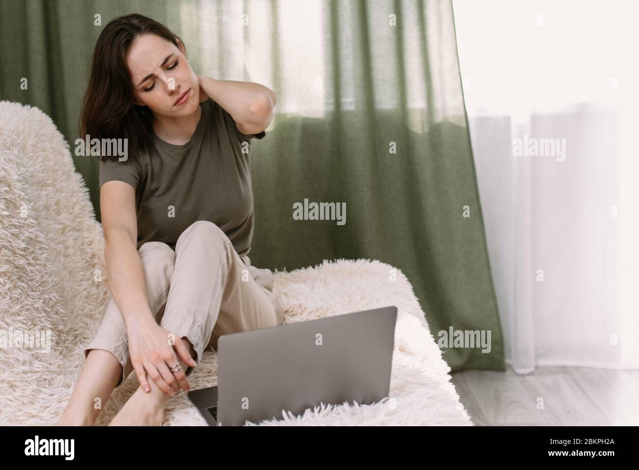 A business woman suffering from pain in neck, stretching up. Tired woman massaging rubbing stiff sore neck tensed muscles fatigue from computer work. Freelancer tired of working at home on the couch. Stock Photo