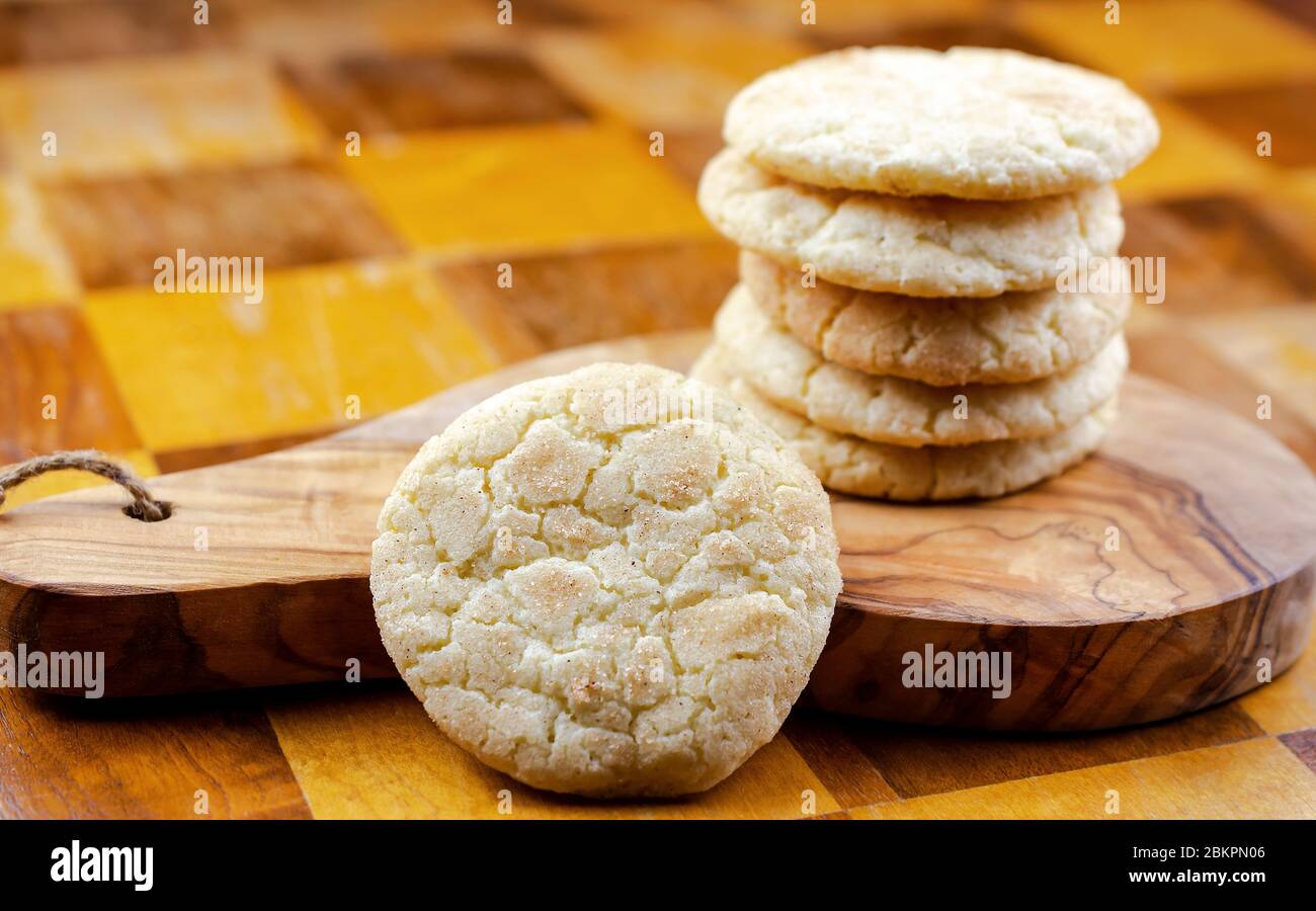 A stack of snickerdoodle cookies on a wooden board sitting on a wooden table.  Landscaped cropped.  Stack of cookies blurred in background.  Focus on Stock Photo