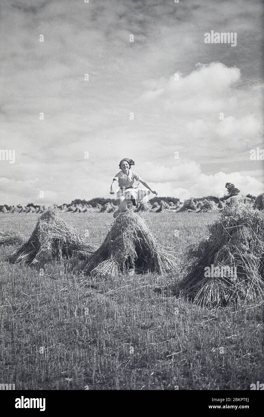 1930s, historical, outside in a recently mowed corn field on a farm, an active young girl jumping a small triangular haystack, Chagford, Devon, England, UK. Stock Photo