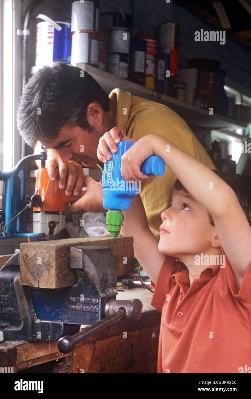Young boy 4-6 years with his father in home DIY shed, copying his father and drilling into a wood block with his play drill. Father and son together, growing, learning, bonding, parenting, nurturing Stock Photo