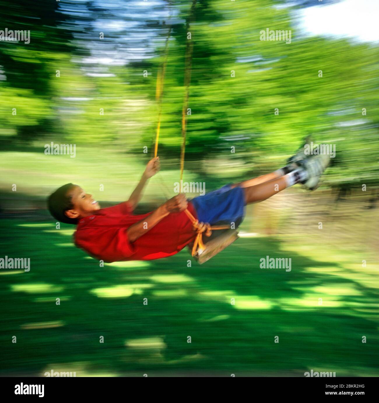 Teenage African American boy 13-15 years growth, energy, summer garden speed blur happily swinging on a rustic rope garden swing, with lush green grass and foliage Stock Photo
