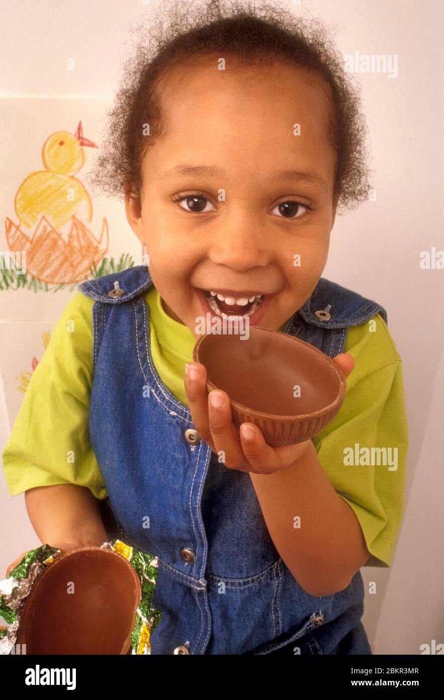 EASTER Infant African American Carribbean 4 years old girl biting into a chocolate Easter egg shell, with her Easter chick drawing on her wall behind Stock Photo
