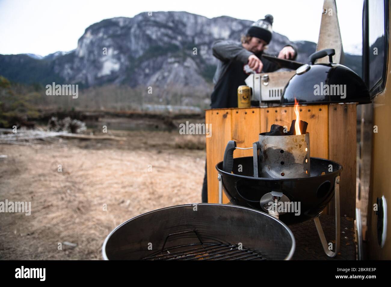 A man cooking on a barbeque set  up at the back of the campervan in a wintry landscape. Stock Photo