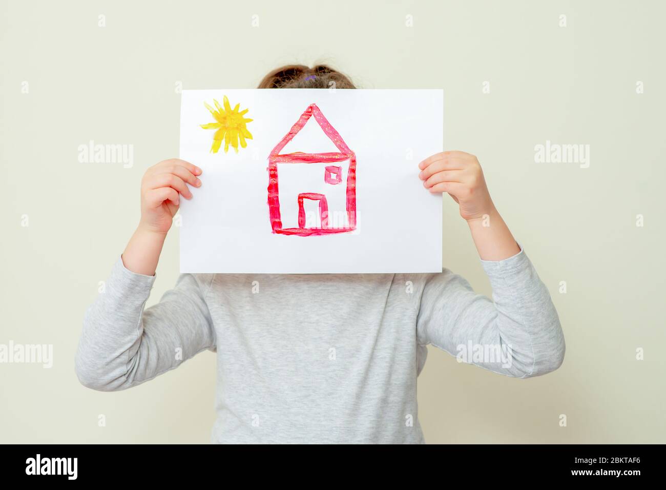 Child is holding picture of house covering her face on yellow background. Children's creativity. Stock Photo