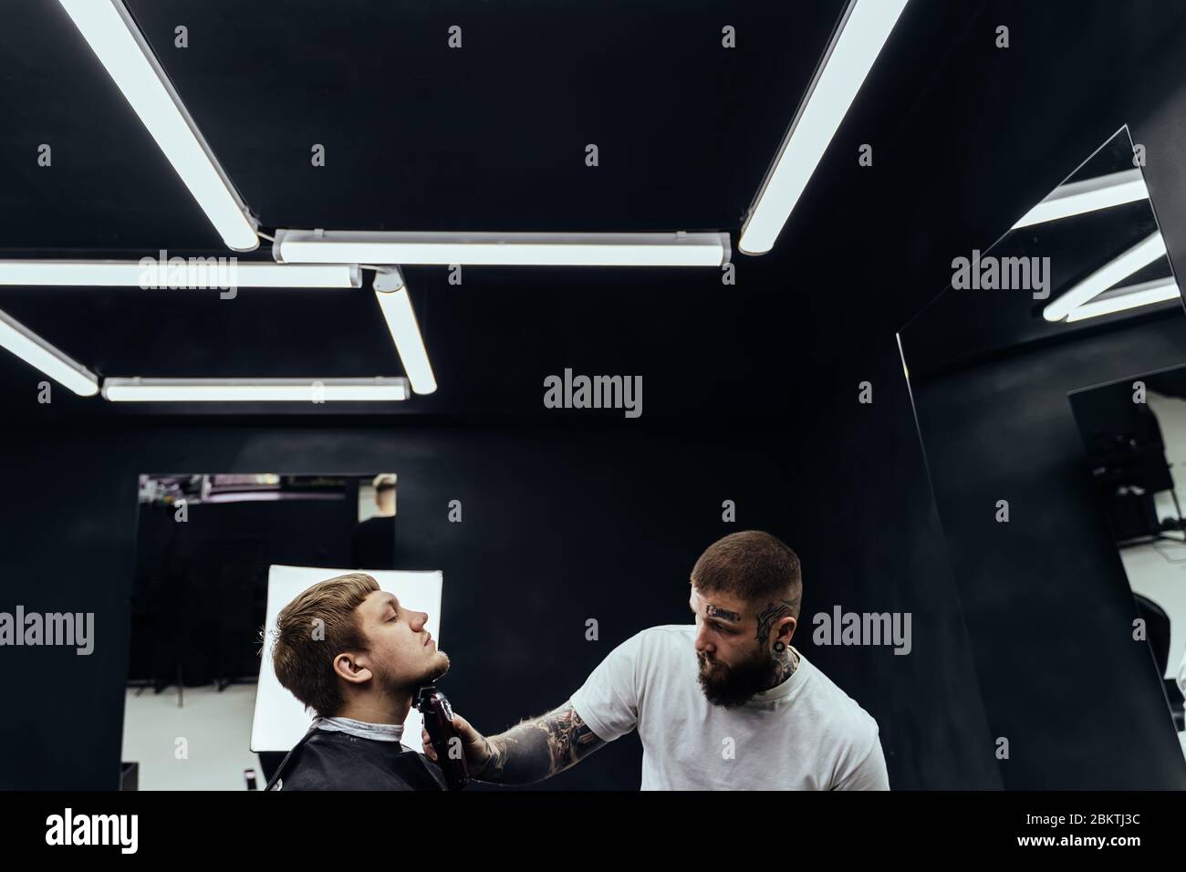 Tattooed Barber trimming bearded man with shaving machine in barbershop. Hairstyling process. Hairstylist cutting the beard of a bearded male. Stock Photo