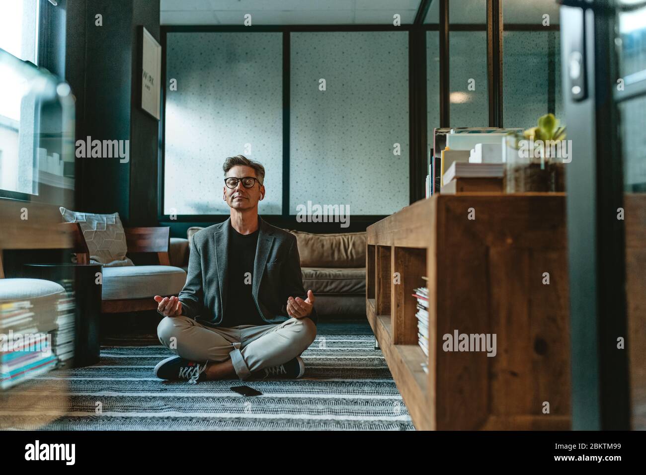 businessman meditating in lotus pose on the floor in the office. Mature business professional doing yoga meditation in office lounge. Stock Photo