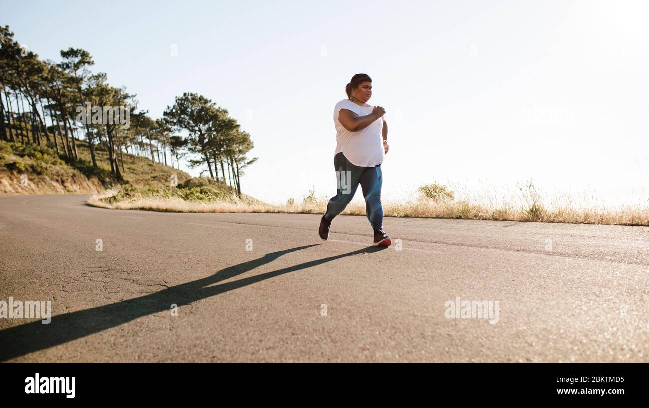 Healthy woman running in morning on a highway. Overweight female jogging outdoors in morning. Stock Photo