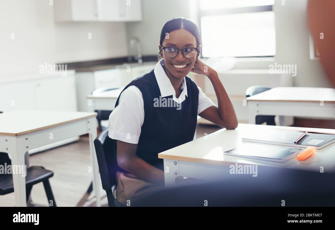 Female student sitting at desk in classroom looking away and smiling. School girl in uniform sitting at her desk with books. Stock Photo