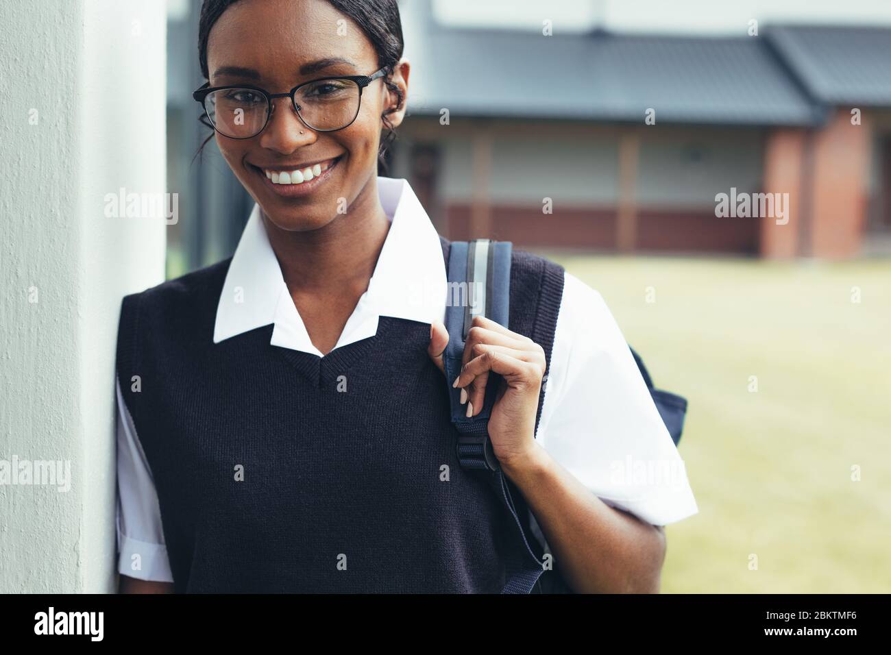 Teenage girl with schoolbag walking through the high school corridor. Female high school student in uniform looking at camera and smiling. Stock Photo