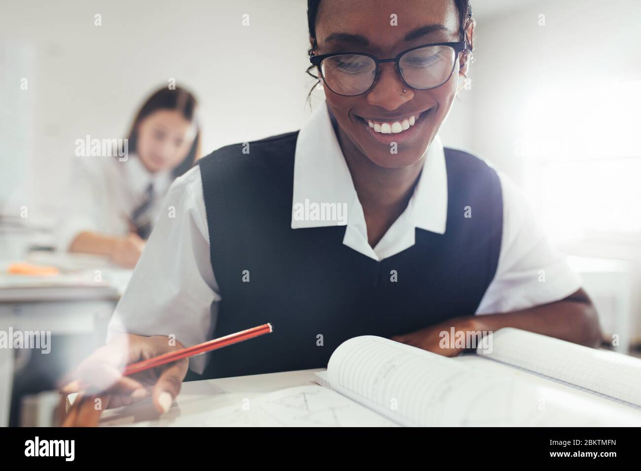 Teenage girl wearing eyeglasses sitting at desk in classroom in high school. Female student smiling while studying in classroom. Stock Photo