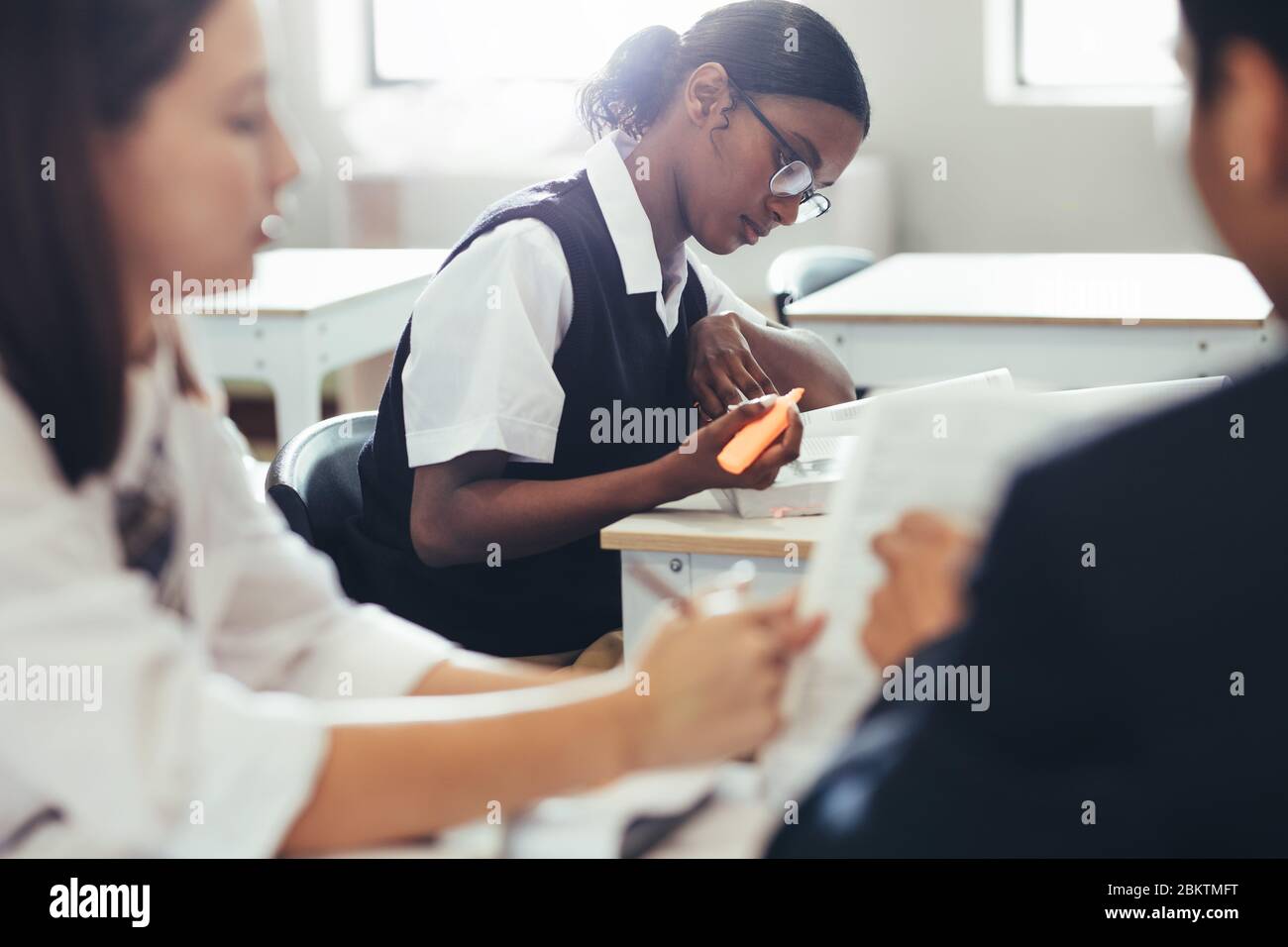 High school students wearing uniform sitting at desk in classroom. Female student studying in classroom with classmate discussing in front. Stock Photo
