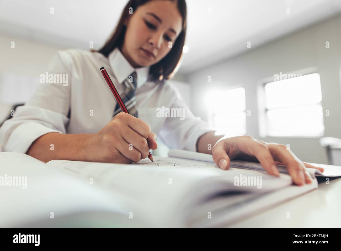 Female student in uniform writing in a book while sitting at desk in high school classroom. Girl student concentrating while writing in a book. Stock Photo