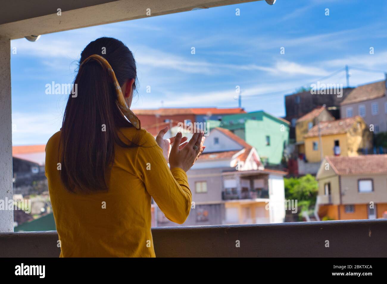 Young woman clapping her hands on the balcony to showi gratitude to all healthcare workers during the coronavirus outbreak. Lots of colorful houses. Stock Photo
