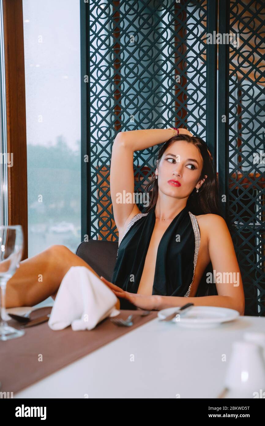 Pretty female with long dark hair is wearing an elegant black evening dress and high heels. She is sitting at a table (for a meal), looking aside. Stock Photo