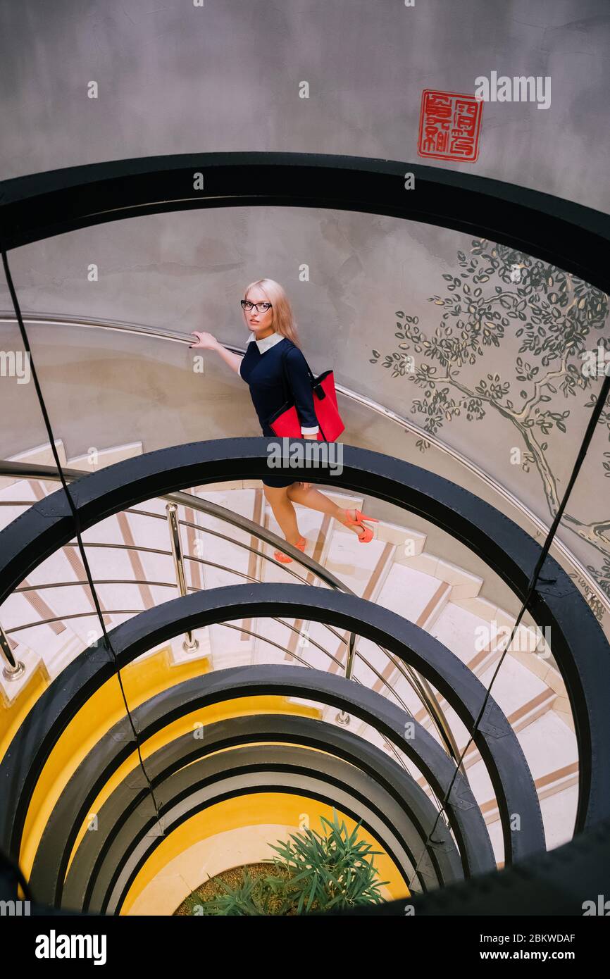 Pretty blonde girl is wearing a formal blue dress, red high heels and glasses; she is going up the stairs and carries a handbag. Stock Photo