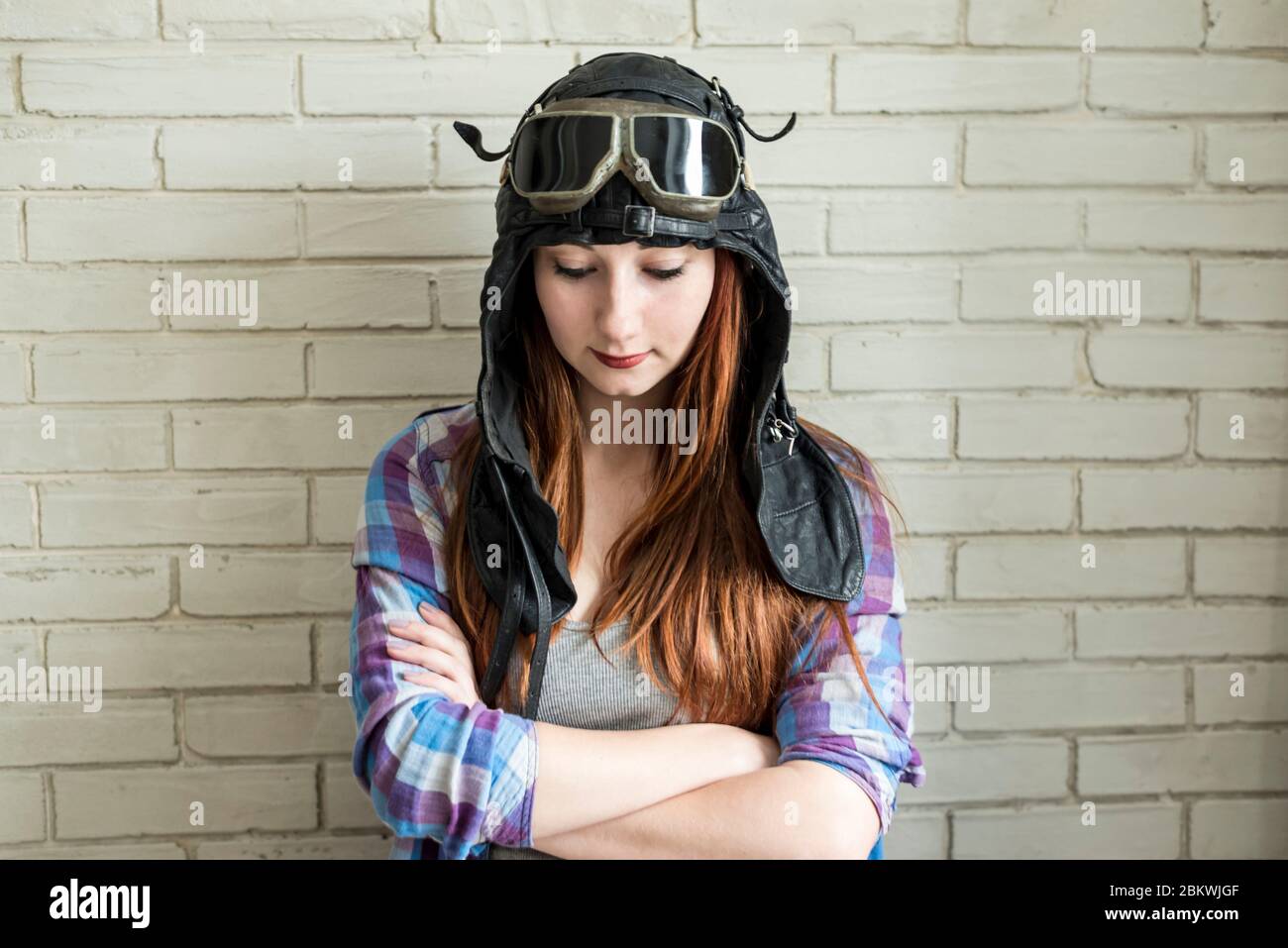 Portrait of a red-haired girl in a pilot's cap and glasses on a brick wall background Stock Photo