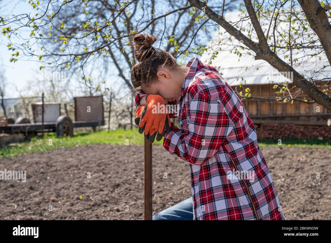 Tired brunette girl in orange work gloves leaned on shovel, resting while working hard in the garden Stock Photo