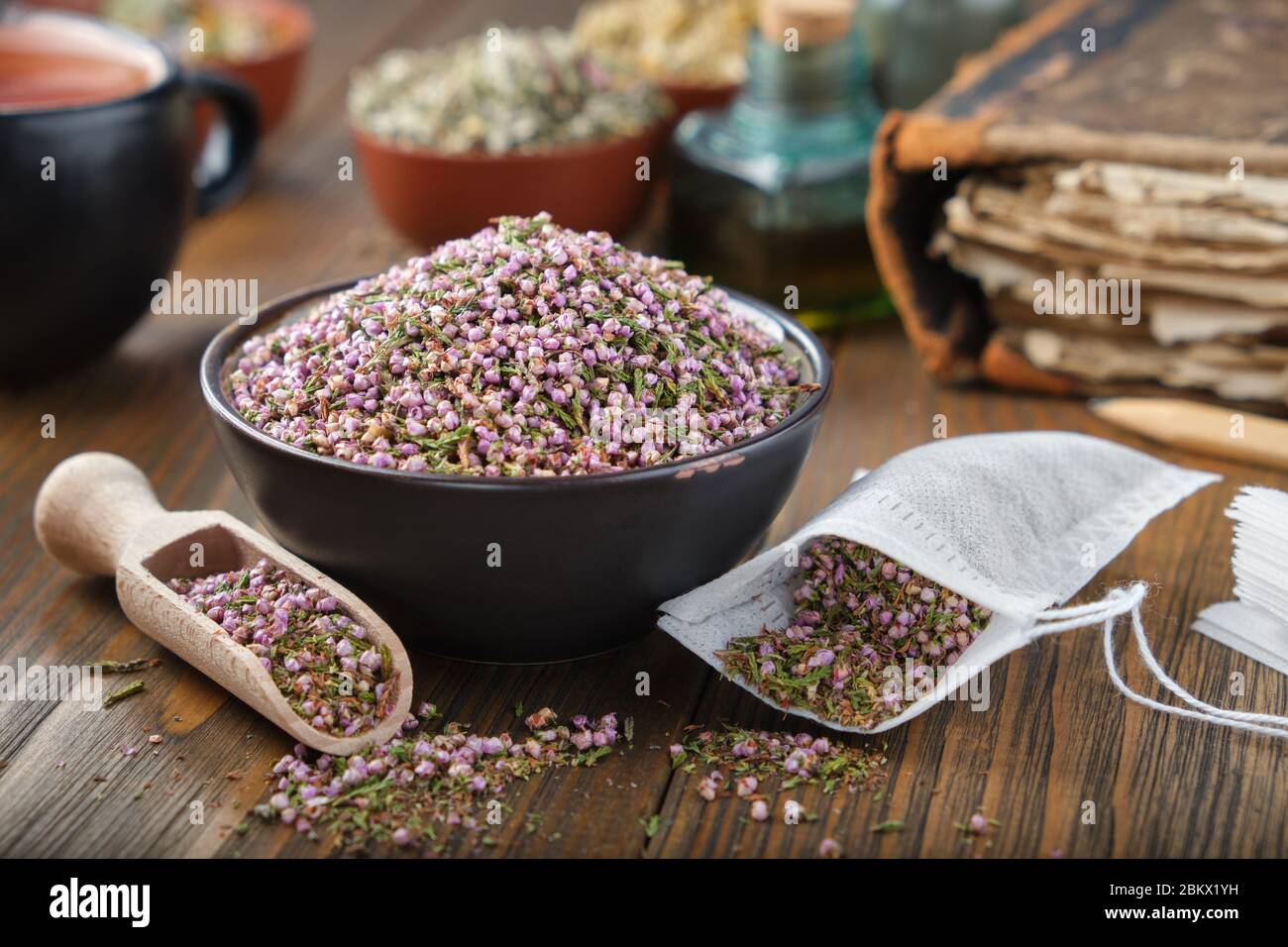 Bowl of dry healthy heather, tea bag with Erica flowers inside. Tea cup, infusion bottle and books on background. Stock Photo