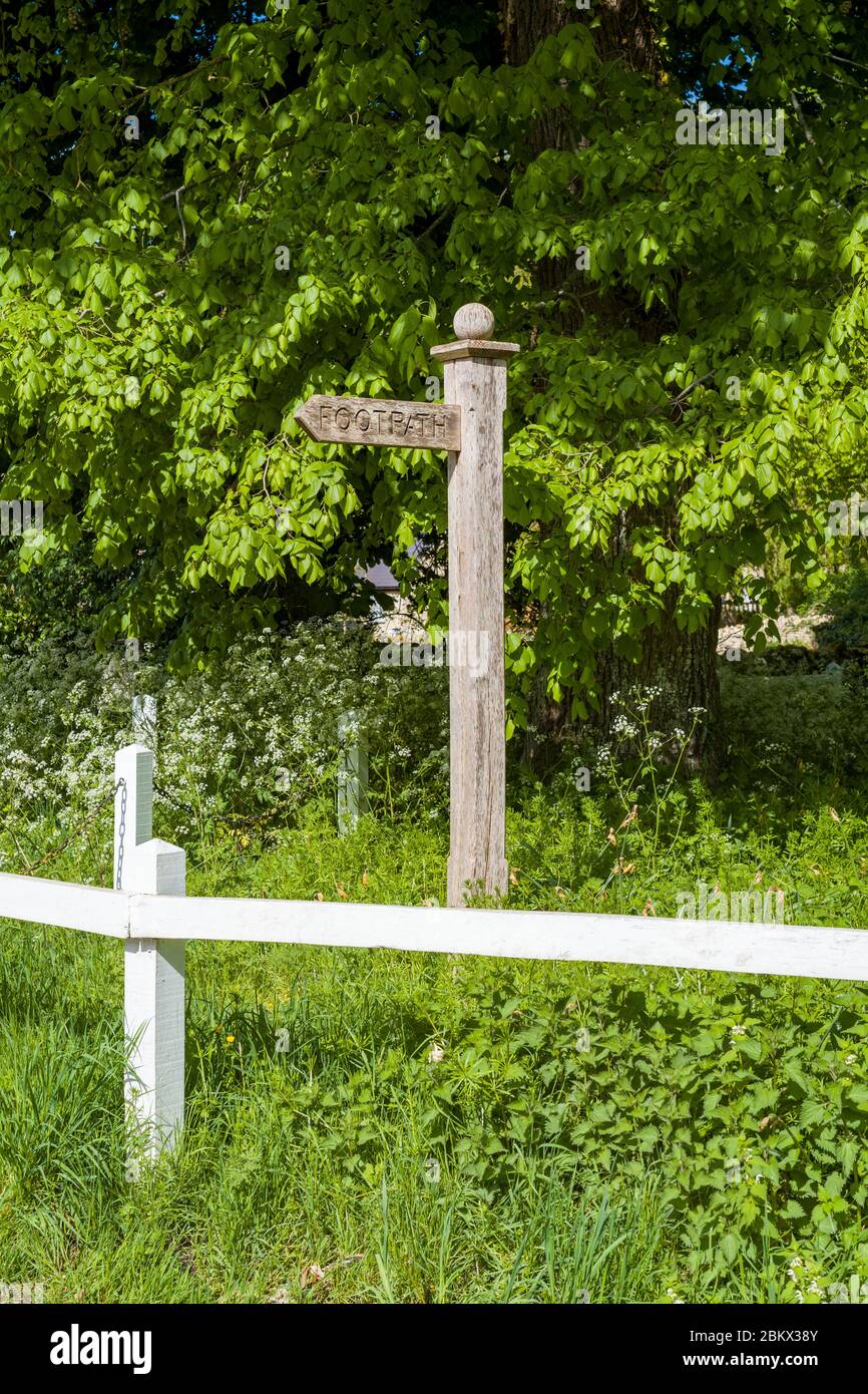 Wooden footpath sign directing walkers to a public footpath and white wooden fence rail in Swinbrook, the Cotswolds, Oxfordshire, UK Stock Photo