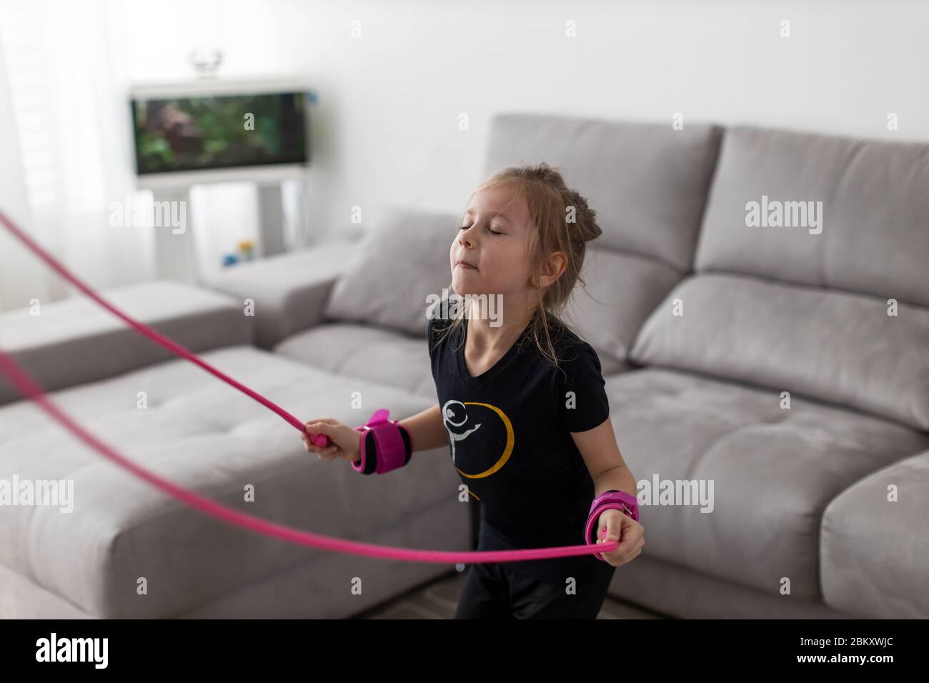 Happy little girl jumping over the rope at home during the isolation. The bright sun illuminates the room Stock Photo